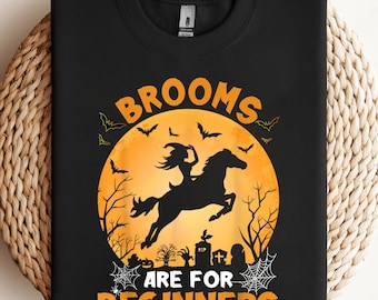 May include: Black sweatshirt featuring a Halloween design. The graphic includes a witch riding a horse in front of a full moon, with bats, trees, and the text "BROOMS ARE FOR BEGINNERS" in orange.