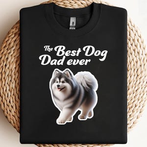 May include: Black sweatshirt with the text "The Best Dog Dad ever" in white script. A graphic of a fluffy, gray and white dog is below the text. The sweatshirt is folded on a woven surface.