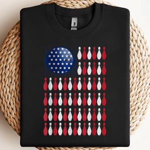 May include: Black sweatshirt featuring a graphic of the American flag, with a bowling ball in place of the stars and bowling pins replacing the stripes. The bowling ball is blue with white stars, and the pins are red and white.