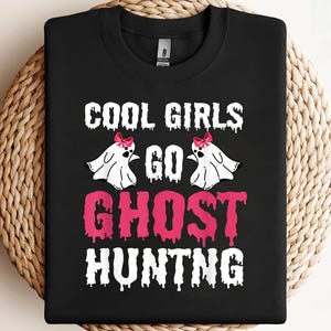May include: Black sweatshirt featuring the text "COOL GIRLS GO GHOST HUNTING" in white and pink. The design includes two white ghost figures with pink bows. The sweatshirt is displayed on a woven surface.
