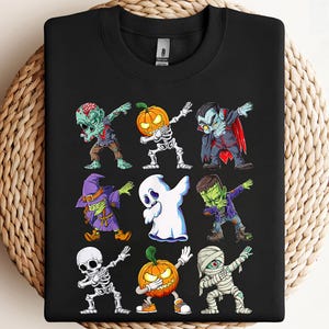 May include: Black sweatshirt featuring a colorful Halloween-themed graphic. The design includes cartoon illustrations of a zombie, skeleton, vampire, witch, ghost, Frankenstein, and mummy, all in a dabbing pose. The artwork is vibrant and playful, perfect for the spooky season.