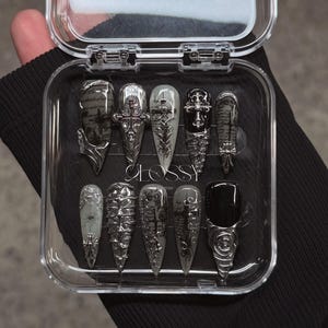 Pode incluir: Um conjunto de dez unhas postiças em estilo gótico numa caixa de plástico transparente. As unhas apresentam enfeites prateados, cruzes e desenhos intrincados. Algumas unhas são pretas sólidas, enquanto outras têm um gradiente cinza ou padrões impressos.