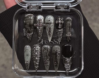 Könnte beinhalten: Ein Set aus zehn falschen Nägeln im gotischen Stil in einer transparenten Kunststoffbox. Die Nägel weisen silberfarbene Verzierungen, Kreuze und komplizierte Designs auf. Einige Nägel sind einfarbig schwarz, andere haben einen grauen Farbverlauf oder gedruckte Muster.