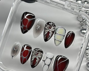 May include: A set of ten almond-shaped press-on nails in a clear plastic case. The designs feature deep red and white base colors with metallic silver accents, including raised butterfly charms, web-like patterns, and thin line art details.