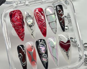 May include: A set of ten press-on nails in a clear plastic case, featuring stiletto shapes with intricate 3D silver metallic accents, swirling red and black marbled patterns, iridescent finishes, and a prominent red heart design on one nail.