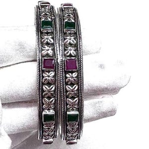 May include: A pair of silver bangles featuring alternating green and red square gemstones. The bangles are decorated with a floral pattern and a textured edge. The jewellery is shown on a white background.