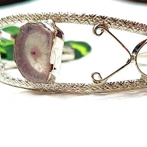 May include: A silver-toned cuff bracelet with a natural stone centerpiece. The stone is a light purple with a white and pink center. The bracelet has a textured band and decorative metalwork.