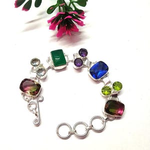 Könnte beinhalten: Ein silbernes Armband mit einer Vielzahl farbiger Edelsteine. Das Armband enthält einen großen, rechteckigen blauen Stein, grüne, lila und rosa Steine. Das Armband wird auf einer weißen Oberfläche mit einer rosa Blume im Hintergrund präsentiert.