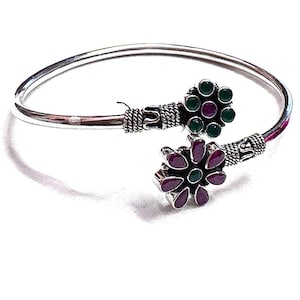 May include: A silver-toned cuff bracelet with floral accents. Each flower features a cluster of dark red and green gemstones. The bracelet has a hinged design for easy wear and a polished finish.