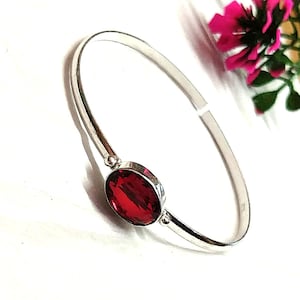 May include: A silver bangle bracelet featuring a large, oval, deep red gemstone. The bracelet has a simple, polished design. The gemstone is set in a silver bezel. The bracelet is displayed on a white surface, with pink flowers in the background.