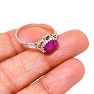 Op de afbeelding: Een zilveren ring met een grote, ovaalvormige, dieproze edelsteen. De edelsteen is gezet in een decoratieve bezel en de ringband heeft een delicaat, vloeiend ontwerp. De ring wordt op een hand getoond.