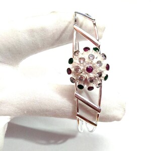 May include: A silver bracelet featuring a central floral design. The floral element is embellished with clear, red, and green gemstones. The bracelet has an open, geometric design with angled bars.
