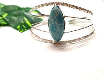 Puede incluir: Un brazalete de puño plateado con una gran piedra turquesa ovalada en el centro. El brazalete tiene un diseño de tres bandas, con patrones texturizados en las bandas superior e inferior. La piedra es de color azul claro con vetas blancas.