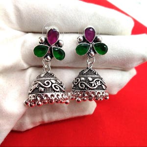 Peut inclure: Une paire de boucles d'oreilles argentées avec un motif floral. Chaque boucle d'oreille présente un groupe de trois pierres vertes et une gemme magenta ovale. Les boucles d'oreilles ont un fond en forme de cloche avec des motifs complexes et de petites perles pendantes.
