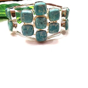 May include: Silver-toned cuff bracelet featuring square, turquoise-colored stones. The bracelet has a double-band design with the stones set in a grid pattern. The stones have a mottled appearance, adding texture to the jewelry.
