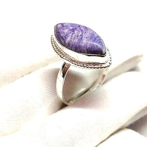 May include: A silver ring featuring a large, oval-shaped purple gemstone. The gemstone has a marbled appearance with white and purple swirls. The ring has a textured border around the stone and a smooth, polished band.