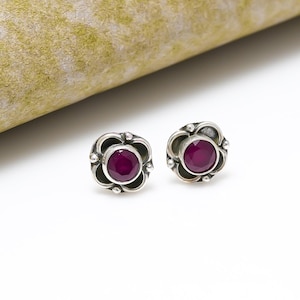 Può includere: Un paio di orecchini a lobo in argento. Ogni orecchino presenta un design a forma di fiore con una gemma rotonda di colore rosso intenso al centro. Gli orecchini sono incastonati in una montatura d'argento decorativa.