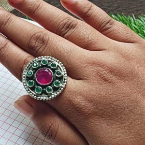 Può includere: Un anello color argento con un design circolare. L'anello presenta una grande pietra rotonda color magenta al centro, circondata da pietre verdi più piccole e un bordo perlato. L'anello è mostrato su una mano.