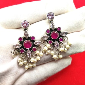 Pode incluir: Um par de brincos prateados com um design floral. Cada brinco apresenta uma pedra grande e redonda de cor magenta no centro, rodeada por pedras magenta menores, cristais transparentes e pérolas brancas penduradas. Os brincos são exibidos em uma superfície branca.