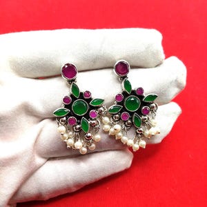 May include: A pair of silver earrings with a floral design. Each earring has a large green centre stone, surrounded by smaller pink and green stones. Small white pearls dangle from the bottom. The earrings are displayed on a white surface against a red background.