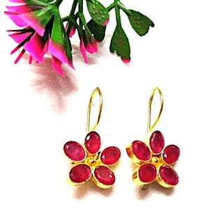 Puede incluir: Un par de pendientes dorados con forma de flor, cada uno con cinco piedras preciosas ovaladas de color rojo rubí. Los pendientes tienen un cierre de gancho. Acentos florales rosas y verdes en el fondo.