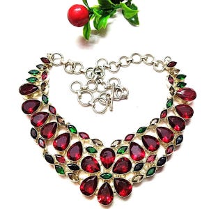 Op de afbeelding: Een zilverkleurige ketting met grote, druppelvormige rode edelstenen. De ketting is versierd met kleinere groene, zwarte en bruine accentstenen. Het heeft een tuimelsluiting en een hartvormig ontwerp.