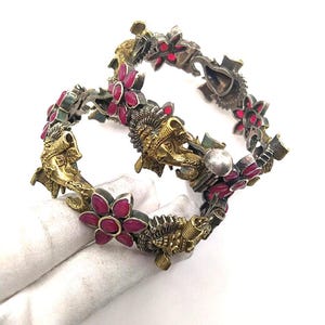 May include: Two ornate silver and gold bangles with red flower accents. The bangles feature detailed designs of mythical creatures and floral patterns. The jewellery is displayed on a white background.