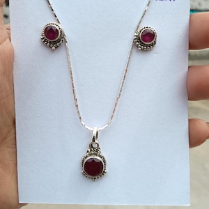 Op de afbeelding: Een zilveren ketting- en oorbellen set met een ronde, dieprode edelsteen hanger en bijpassende oorstekers. De sieraden hebben een gedetailleerd, sierlijk ontwerp rond de edelstenen. De set wordt gepresenteerd op een witte kaart.