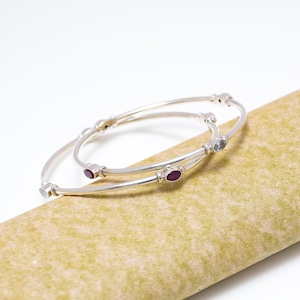May include: Two silver bangles with oval-shaped gemstones in shades of red and light blue. The bangles are resting on a textured, tan surface. The jewelry is displayed against a white background.