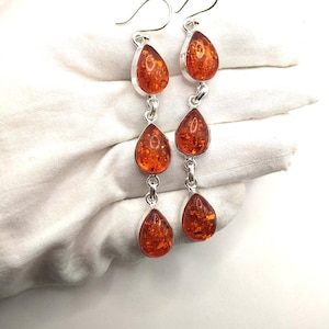 May include: Dangle earrings with three amber teardrops linked by silver-coloured metal. The amber stones, with a warm orange-brown hue, are set in silver-coloured bezels. The earrings have a hook closure. The earrings are approximately 7 cm in length.