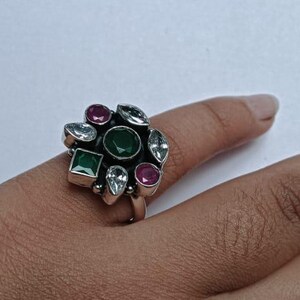 May include: A silver ring with a cluster of colorful gemstones. The ring features a large, oval-shaped green stone at the center, surrounded by smaller gems in shades of red, clear, and green. The ring is worn on a finger.