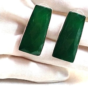 Puede incluir: Un par de pendientes rectangulares de piedra preciosa verde con ribete de metal plateado. Los pendientes son de color verde esmeralda vibrante y tienen una superficie facetada. Los pendientes se muestran sobre una superficie blanca.