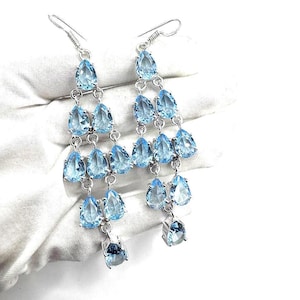 Puede incluir: Un par de pendientes de araña con múltiples niveles de piedras preciosas en forma de lágrima de color azul claro. Los pendientes están suspendidos de ganchos y cadenas de color plateado. Las piedras preciosas están facetadas, reflejando la luz. Los pendientes se muestran sobre un fondo blanco.