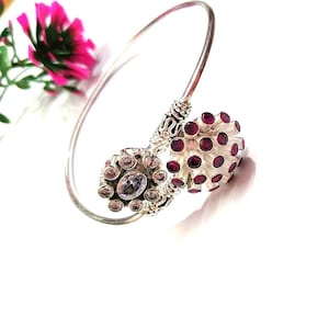 May include: A silver-coloured bracelet with two decorative clusters. One cluster features a central oval-shaped stone surrounded by smaller stones. The other cluster has a textured design with numerous small, round, red stones. The bracelet is set against a white background.