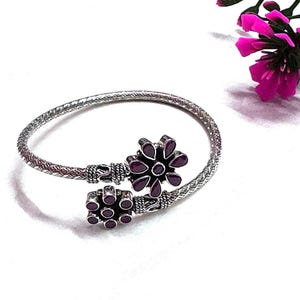 Peut inclure: Bracelet manchette argenté avec deux motifs floraux ornés de pierres rouges foncées. Le bracelet présente un motif tressé texturé et une ouverture à charnière. Une branche de fleurs roses est en arrière-plan.
