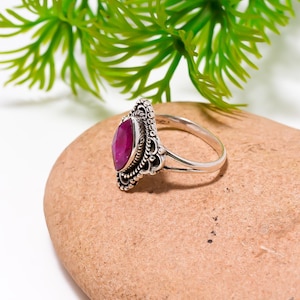 Peut inclure: Une bague en argent ornée d'un grand rubis de forme ovale. La bague présente un sertissage orné et détaillé avec un motif de filigrane autour du rubis. La bague est présentée sur une pierre brune, avec un feuillage vert en arrière-plan.