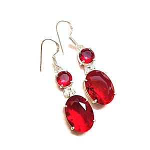 Può includere: Orecchini pendenti con montatura in metallo color argento. Ogni orecchino presenta tre pietre preziose rosse: una piccola pietra rotonda in alto, una piccola pietra rotonda al centro e una grande pietra ovale in basso. Le pietre sono di un rosso vibrante.