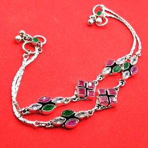 May include: Silver anklet with a red background. The anklet features a delicate chain design with alternating red, green, and clear gemstones. Small bells and green gemstones adorn the ends of the anklet. The gemstones are set in a silver metal.