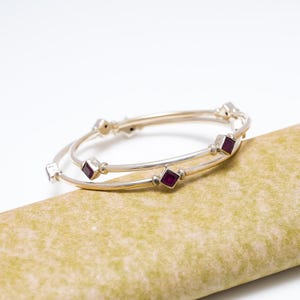 Puede incluir: Dos brazaletes de plata con gemas cuadradas de color rojo intenso. Los brazaletes son delgados y tienen un diseño sencillo, con las gemas espaciadas uniformemente alrededor de la circunferencia. El fondo es de color verde claro.