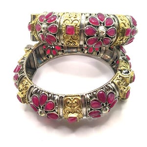 Könnte beinhalten: Zwei verzierte silber- und goldfarbene Armreifen mit leuchtend roten Edelsteinen. Die Armreifen weisen komplizierte Blumen- und geometrische Designs auf, wobei die roten Steine in einer blütenblattartigen Anordnung angeordnet sind. Der Schmuck wird vor weißem Hintergrund präsentiert.