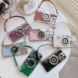 May include: A collection of camera-shaped phone cases in various colors, including brown, pink, green, and black. Each case features a silver-colored lens and the text "W088A". The cases have matching straps.