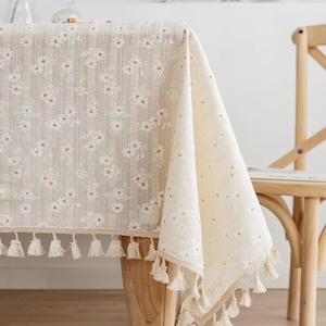May include: Beige tablecloth with a daisy pattern. The tablecloth has a fringe of tassels along the bottom edge. The daisy pattern is white with yellow centres. The tablecloth is draped over a wooden table.