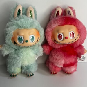 May include: Two plush figures, one mint green and one pink, each with a furry bunny-like hood and a doll-like face. The figures have large eyes, small mouths with braces, and small arms and legs. The ears are decorated with glitter.