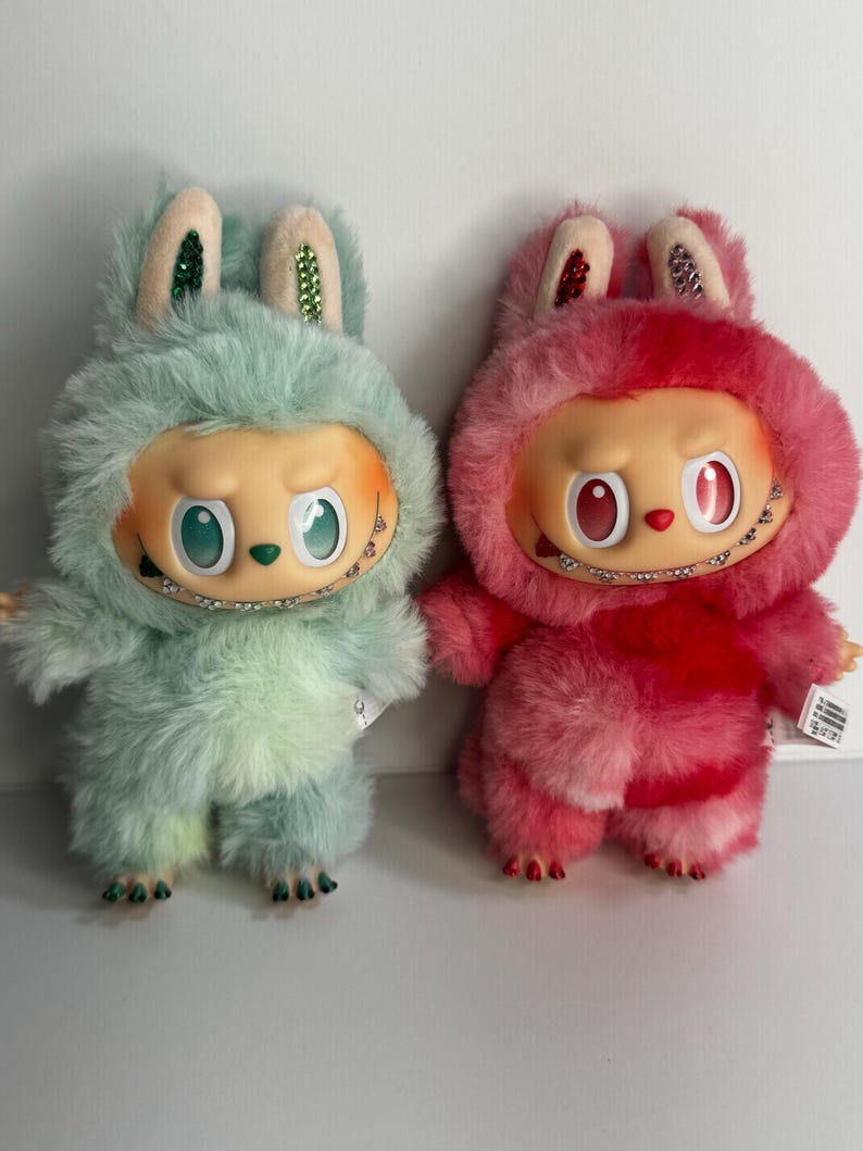 May include: Two plush figures, one mint green and one pink, each with a furry bunny-like hood and a doll-like face. The figures have large eyes, small mouths with braces, and small arms and legs. The ears are decorated with glitter.