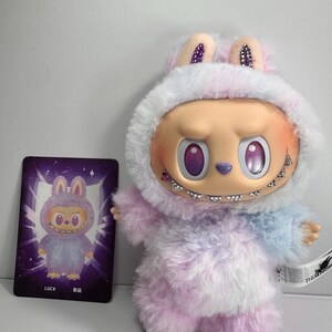 May include: A plush toy figure in a fuzzy, pastel-colored outfit with a hood and bunny ears. The figure has a distinctive face with large, purple eyes and a stitched smile. A card with a matching illustration is next to the toy.