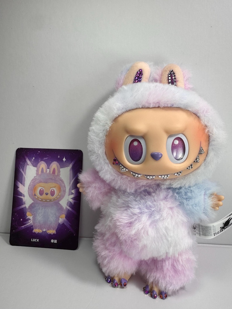 May include: A plush toy figure in a fuzzy, pastel-colored outfit with a hood and bunny ears. The figure has a distinctive face with large, purple eyes and a stitched smile. A card with a matching illustration is next to the toy.
