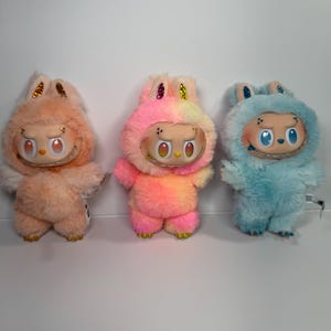 May include: Three plush figures in a row. The figures have a cartoonish design with large eyes and a smiling mouth. The figures are in shades of peach, pink, and blue. Each figure has a fluffy, furry body and a hood with glitter accents.