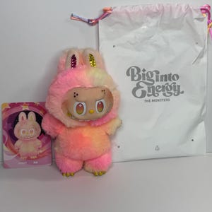 May include: A plush toy in the shape of a bunny with a two-tone pink and yellow fur. The toy has a smiling face with large eyes and a necklace. A card with a bunny illustration and a white bag with "Biginto Energy" text are also present.