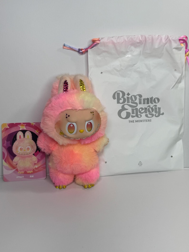 May include: A plush toy in the shape of a bunny with a two-tone pink and yellow fur. The toy has a smiling face with large eyes and a necklace. A card with a bunny illustration and a white bag with "Biginto Energy" text are also present.