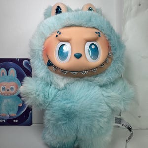 May include: A plush toy in the shape of a creature with a light blue furry body and head covering. The toy has large blue eyes, a smiling mouth with braces, and small ears. A small card with a similar character is visible.
